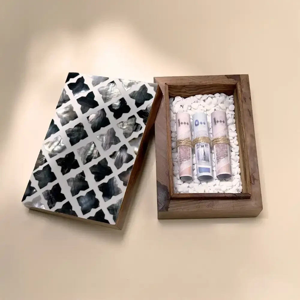 Open wooden money box with mosaic lid beside rolled cash on white pebbles