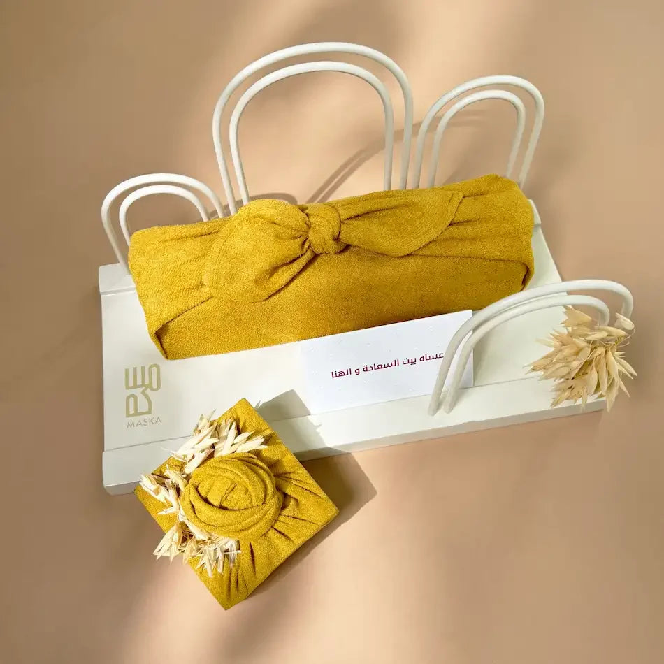 Mustard furoshiki-wrapped parcels beside Maska gift tray, top-down view with greeting card
