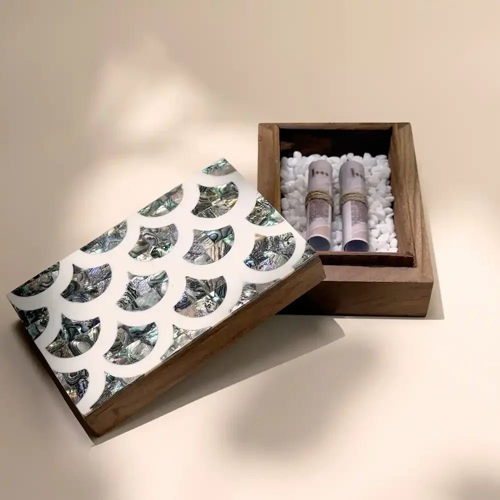Open wooden money box showing rolled cash on white pebbles beside mother-of-pearl lid
