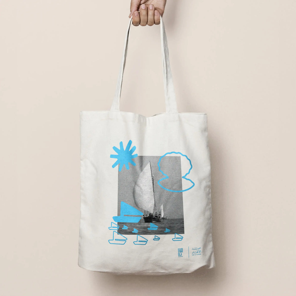 Canvas tote bag with dhow sailboat print in blue, held by hand against beige backdrop