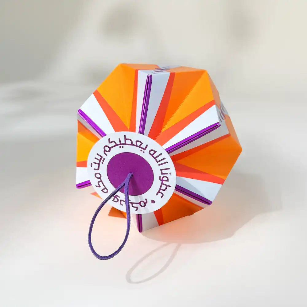 Folded paper lantern resting on its side showing the sunburst pattern and circular Arabic tag