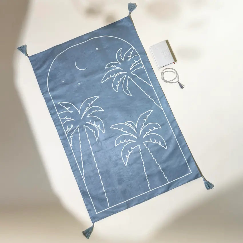 Blue prayer mat with palm tree print, beaded tasbih and Quran beside it