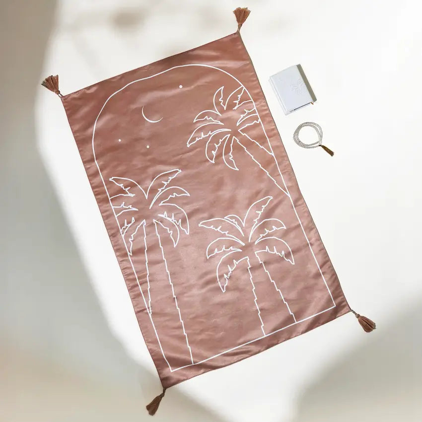 Brown palm tree print prayer mat with tassels beside Quran and prayer beads