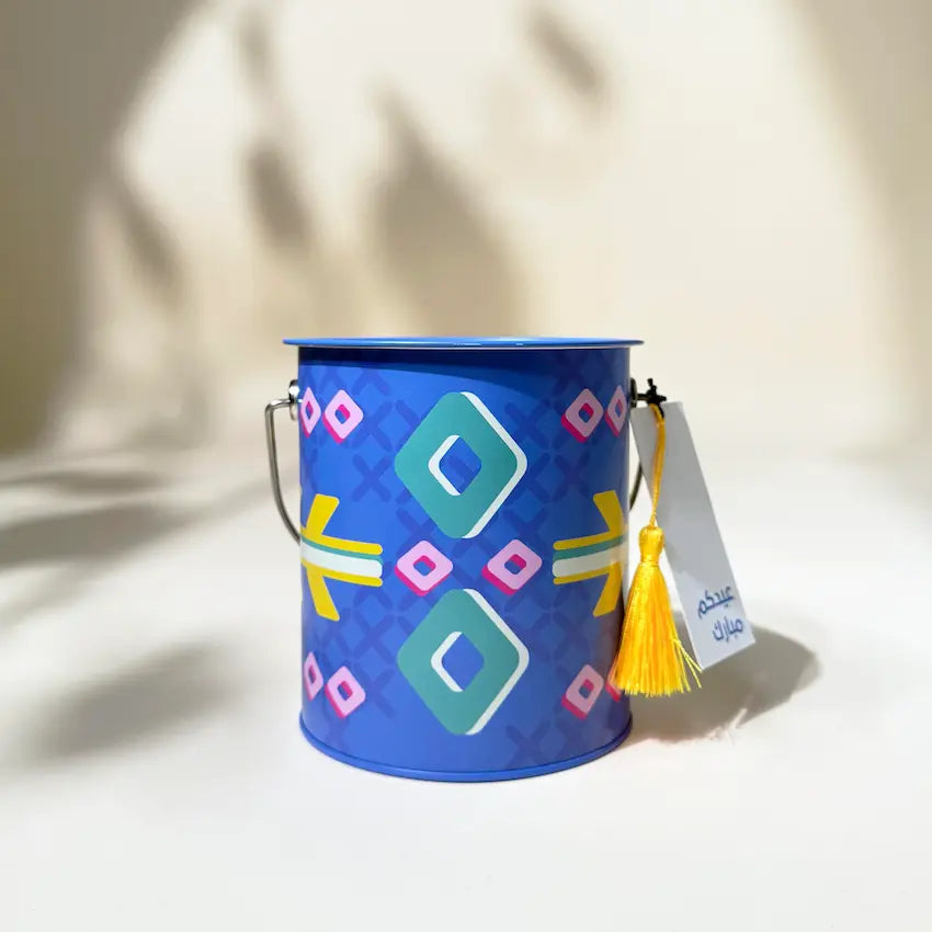 Blue Sadu-patterned metal can with wire handle and yellow tassel tag, front view